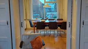A room with a dining area, a window with curtains, a table, and a chair with a cushion. - Authentic dike house Nieuw Vliet (Nieuwvliet)