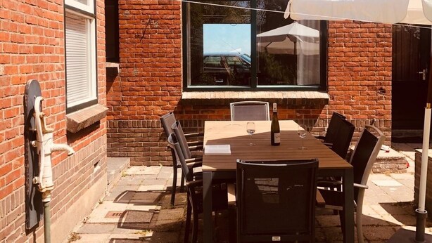 A brick building with a patio area featuring a table and chairs, a white umbrella, and a window with a view of a car. - Authentic dike house Nieuw Vliet (Nieuwvliet)