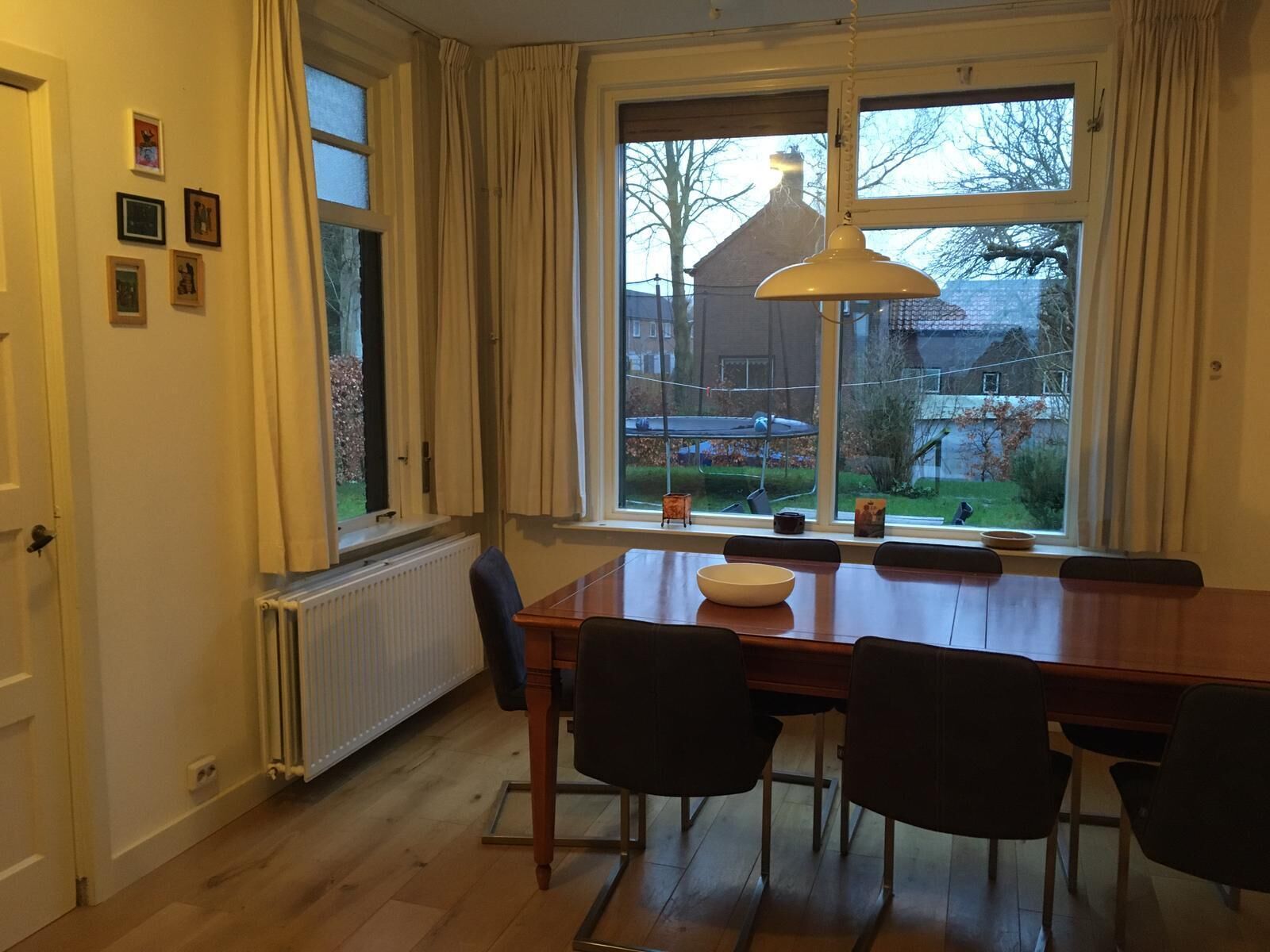 Comedor con mesa de madera, cuatro sillas, lámpara colgante y vista a zona residencial a través de amplios ventanales.