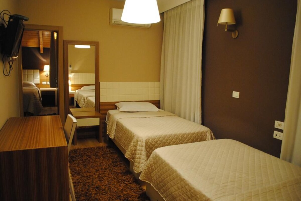 Quarto de hotel com duas camas, uma escrivaninha de madeira, um espelho, uma luminária e piso coberto por carpete.