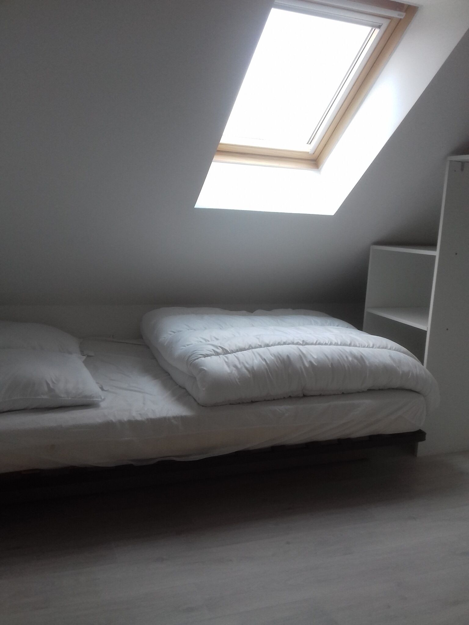 A small, dimly lit room with a single bed, a skylight, and a white wardrobe.