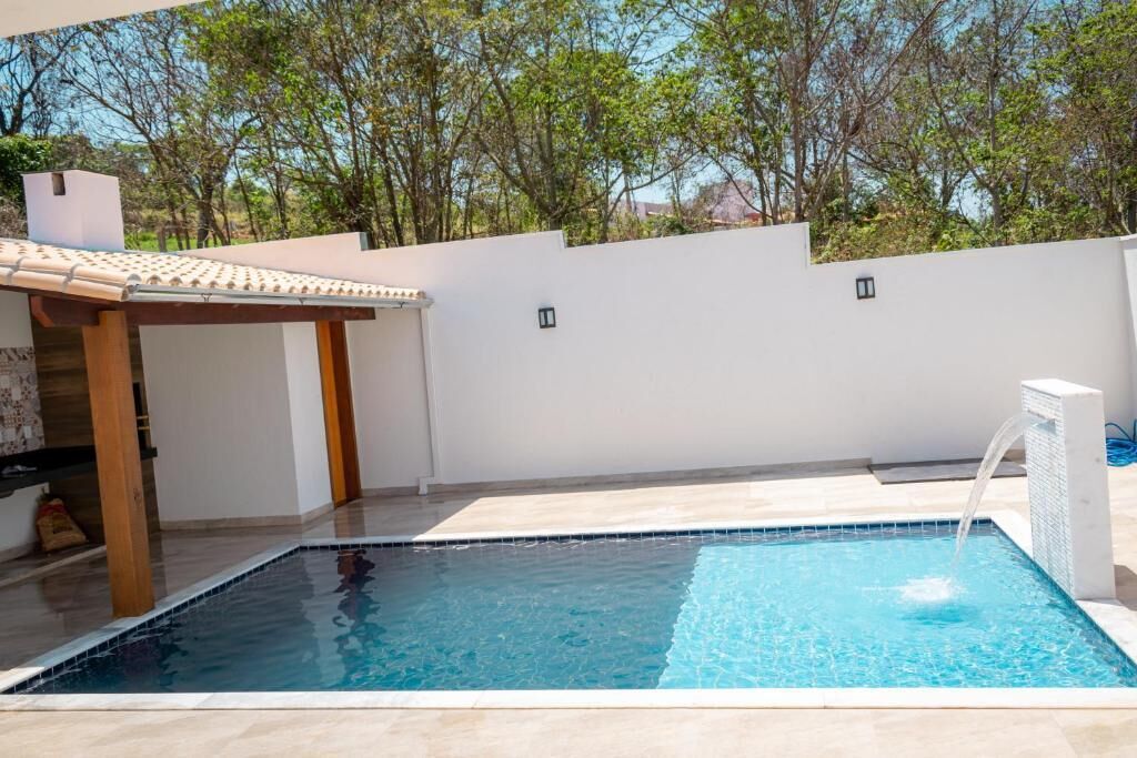 Una piscina con una pérgola y una fuente, rodeada por un muro blanco y árboles.