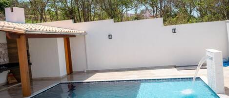 Una piscina con una pérgola y una fuente, rodeada por un muro blanco y árboles.