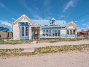 A two-story house with a blue roof and white walls, featuring a front porch and a small garage.