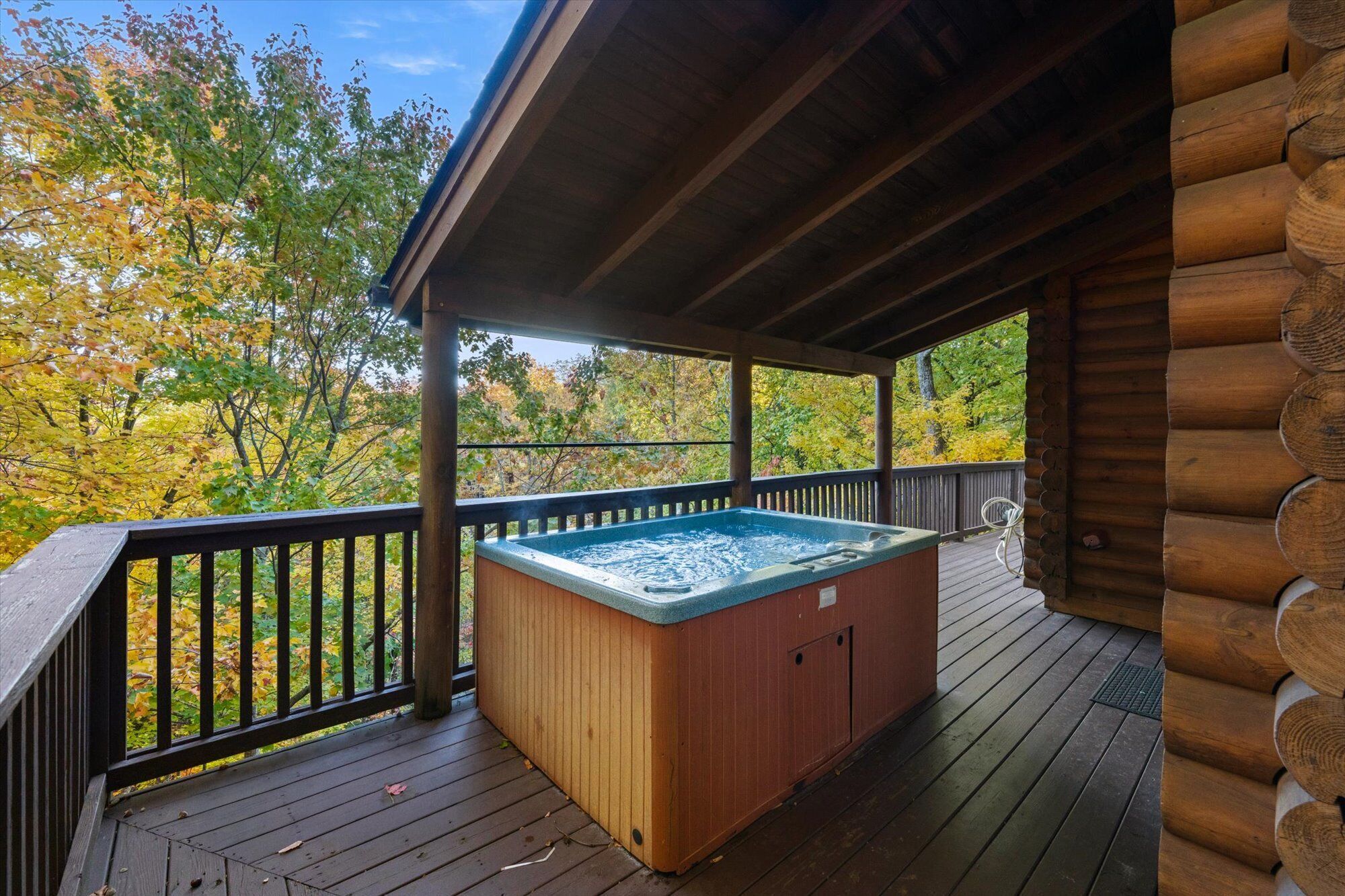 Eine Holzterrasse mit Whirlpool, umgeben von einem Holgitter, mit Blick auf einen Wald im Herbstlaub.