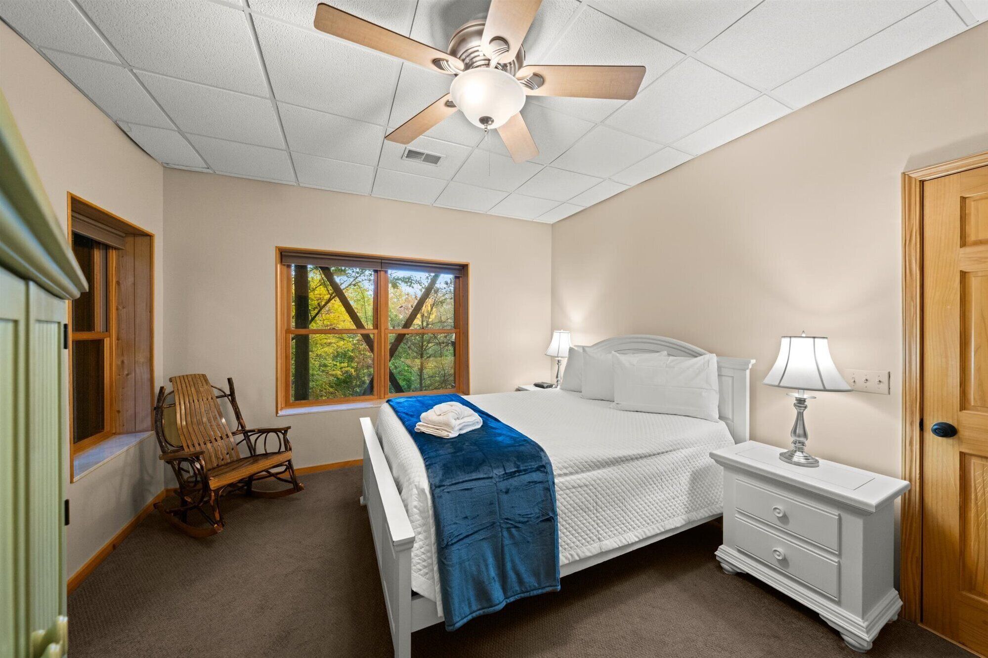A bedroom with a bed, a nightstand, a rocking chair, a window with a view of trees, and a ceiling fan.