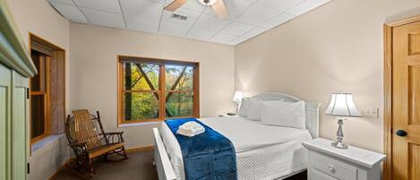 Un dormitorio con cama, mesita de noche, sillón mecedor, ventana con vista a árboles y ventilador de techo.