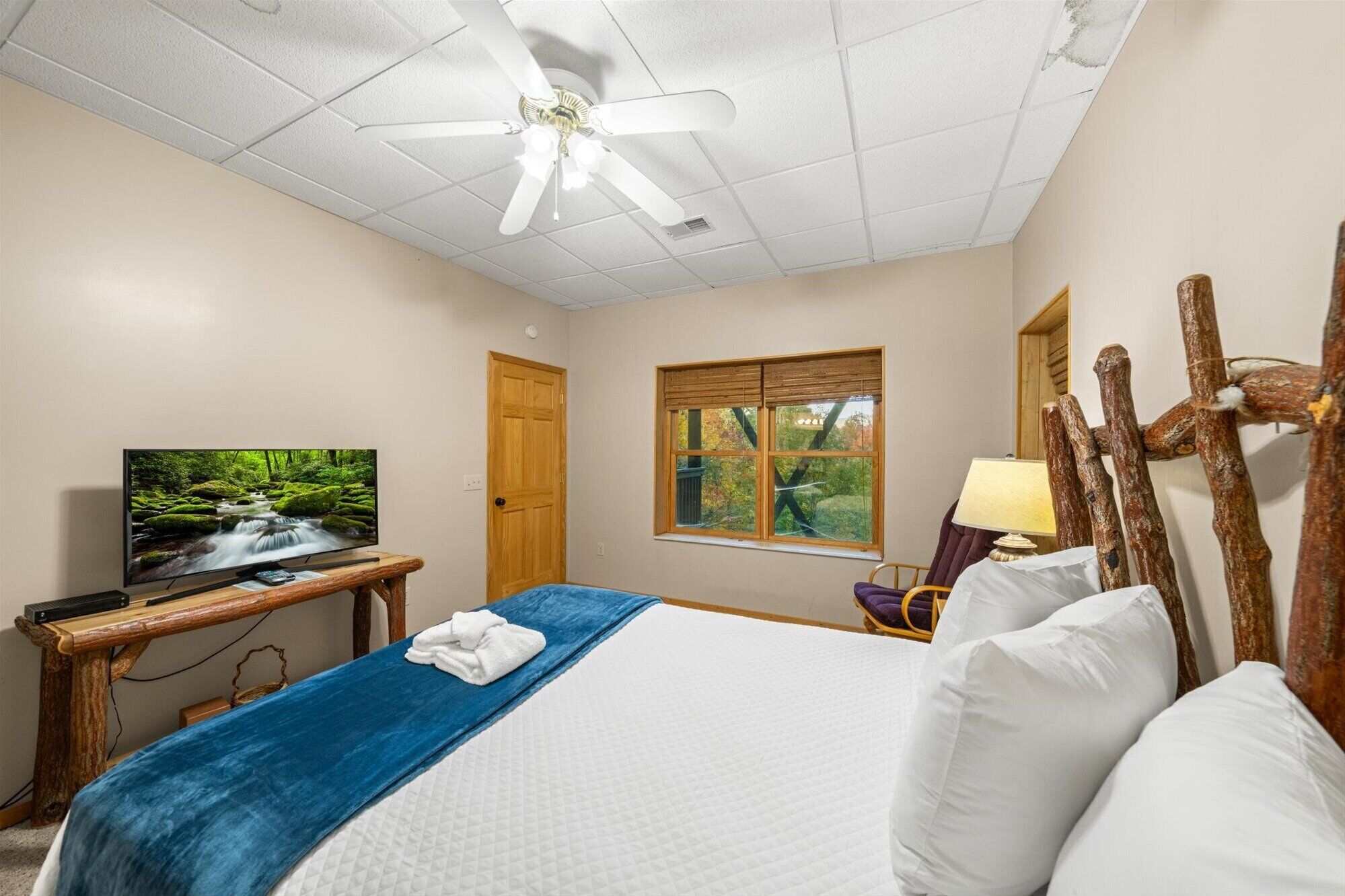 Quarto de hotel com cama, televisão sobre suporte de madeira, janela com vista para árvores e ventoinha no teto.