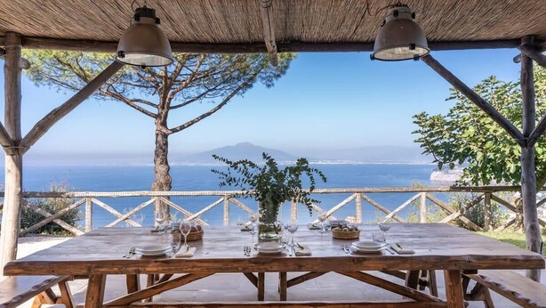 A wooden table set for dining with a view of the sea and a mountain in the background. - Villa Giulia: Views, Pool & Concierge by Feeling Italy (Sorrento)
