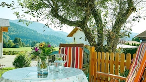 A round table with a floral tablecloth, a pitcher, and glasses, set outdoors with a wooden chair and a striped cushion.