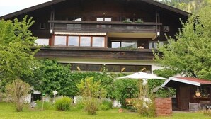 A two-story wooden chalet with balconies, surrounded by lush greenery and a mountain backdrop.