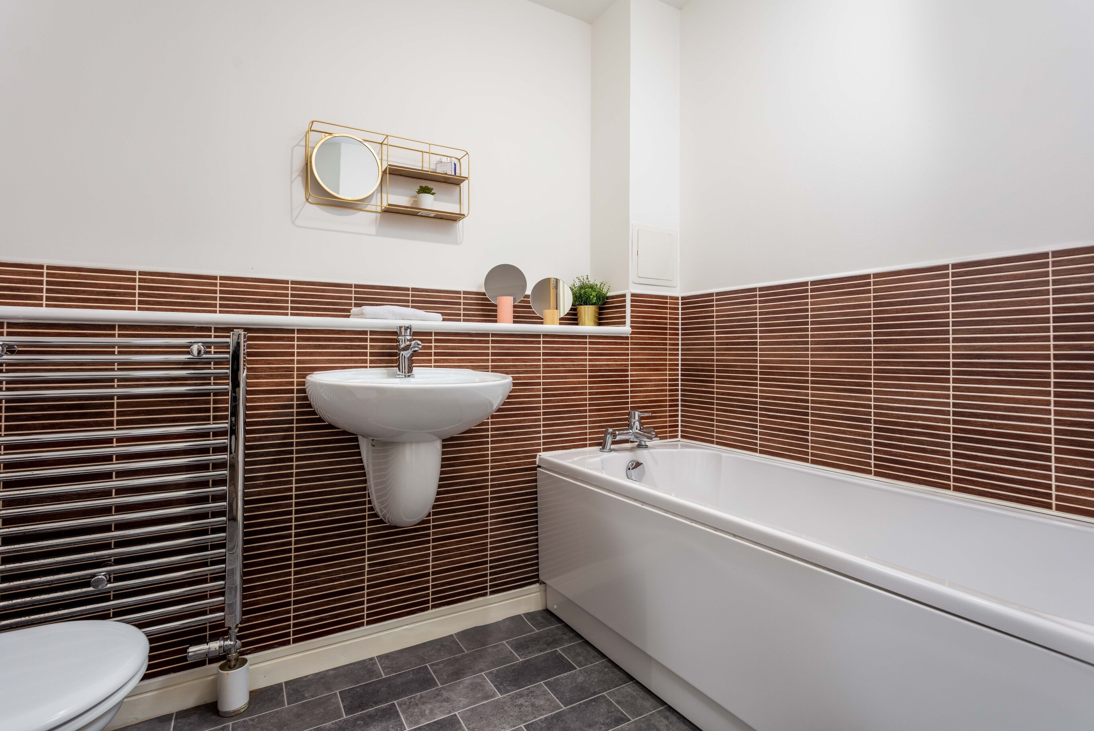 Un baño moderno con una bañera blanca, un lavabo blanco y un inodoro. Hay un perchero y una repisa con objetos decorativos.