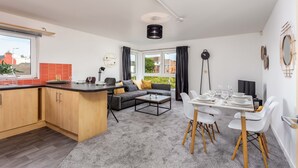 A modern living room with a sofa, coffee table, and dining area. There is a kitchen with wooden cabinets and a window overlooking a garden. - Walker Suite No82 - Donnini Apartments (Kilmarnock)