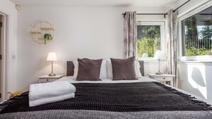 A neatly made bed with a black and gray blanket, two pillows, and a folded white towel. There is a bedside lamp on the left and a window with curtains on the right. - Walker Suite No82 - Donnini Apartments (Kilmarnock)