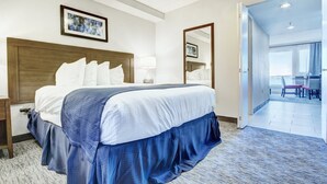 Une chambre d’hôtel avec un grand lit, une tête de lit en bois, une table de chevet, une lampe de chevet, un miroir et une vue sur la salle à manger par une porte ouverte.
