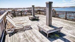 Une terrasse en bois avec des chaises longues et un banc en bois, donnant sur une étendue d’eau avec des bâtiments en arrière-plan.