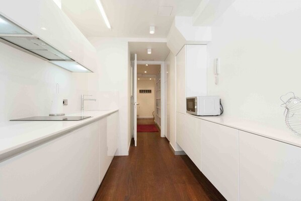 A narrow, modern kitchen with white cabinetry, a microwave, and a red rug leading to a hallway. - Gros Beach | The Rentals Collection (San Sebastián/Donostia)
