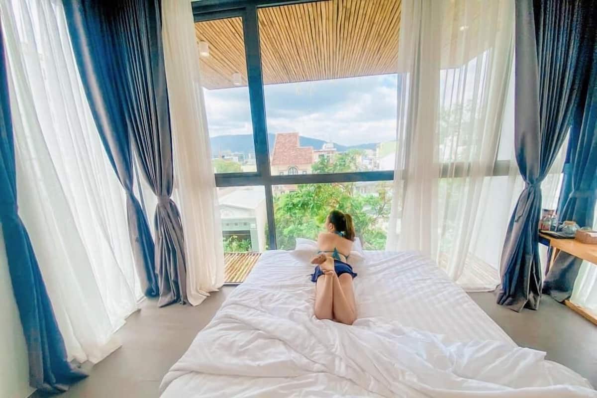 A bedroom with a large window, a bed, and a view of buildings and greenery outside.