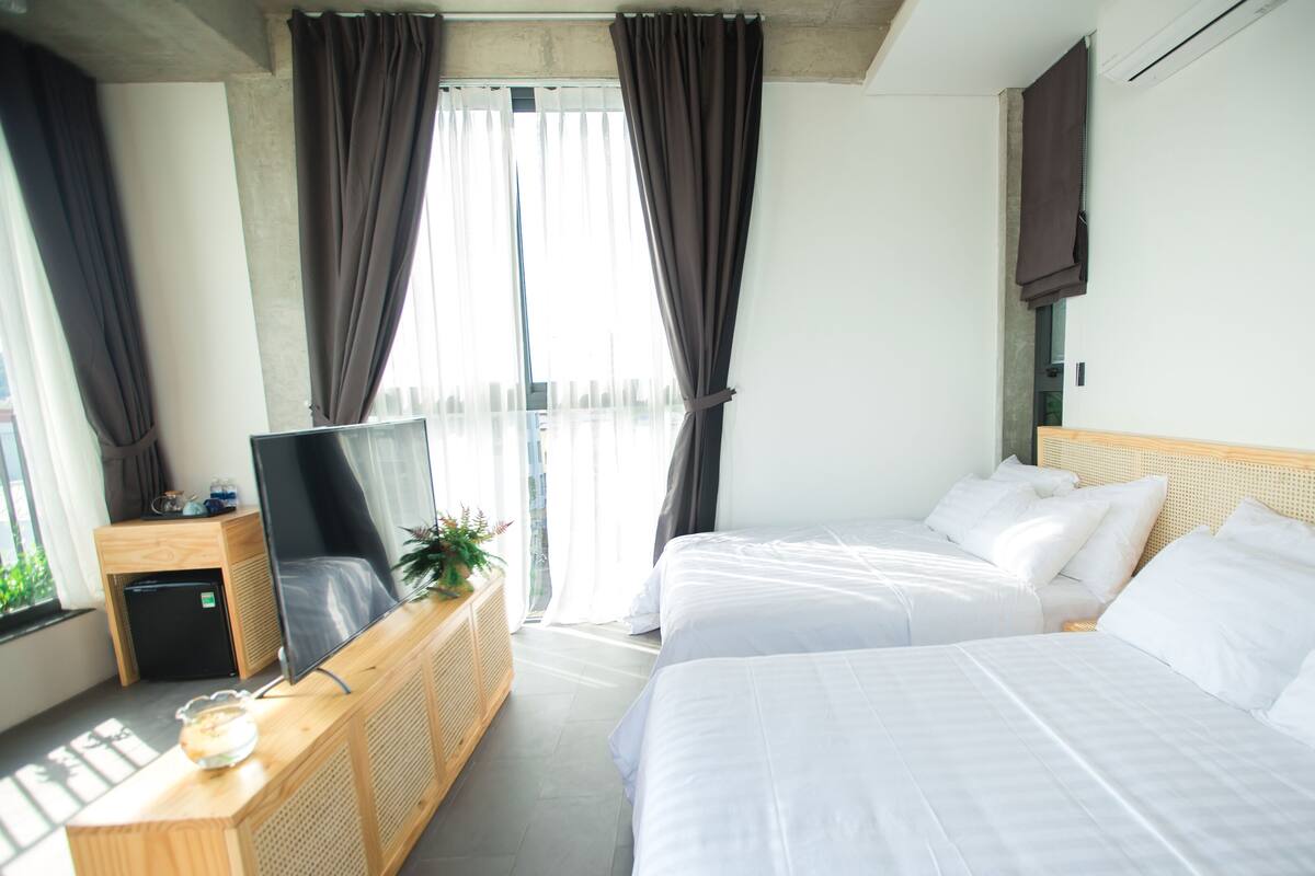 A hotel room with two beds, a TV on a wooden stand, and a view of the outdoors through large windows.