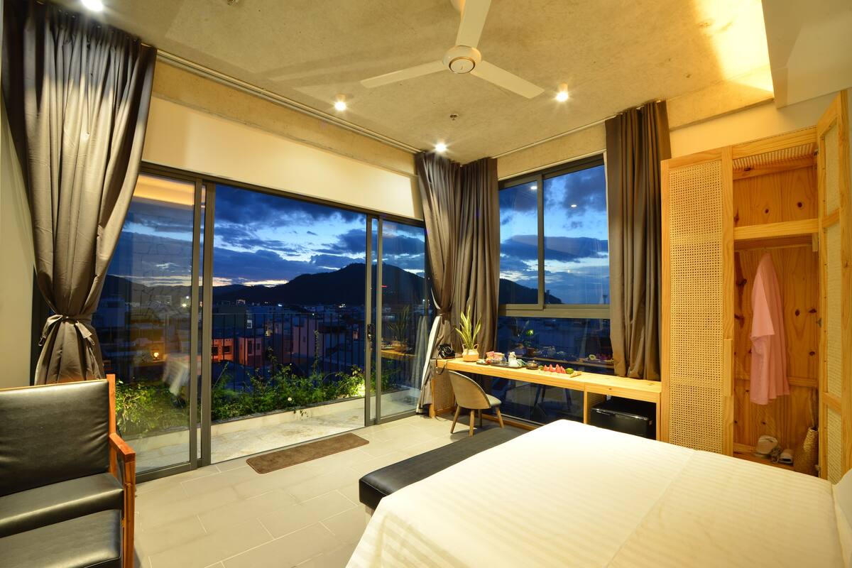 A modern bedroom with a large bed, a desk, and a chair. It features a large window with a view of the city and mountains.
