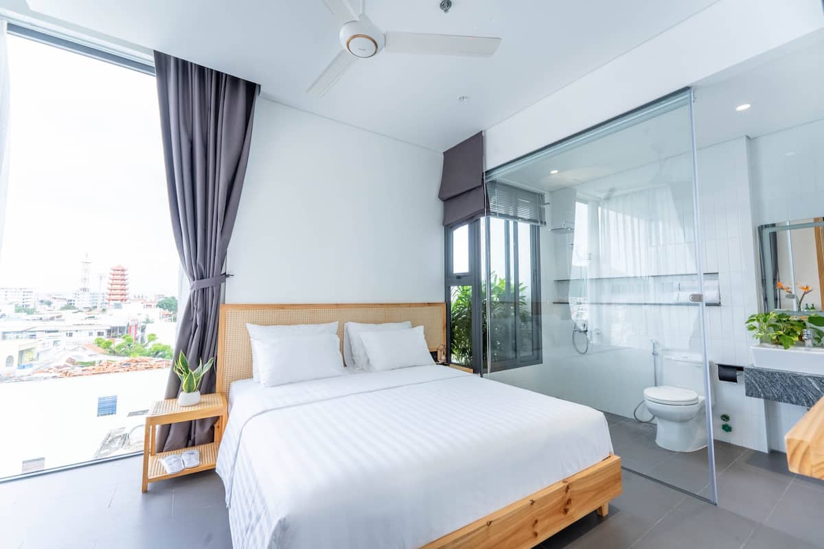 A modern bedroom with a large bed, a bedside table with a plant, a view of the city, a glass shower enclosure, and a toilet.