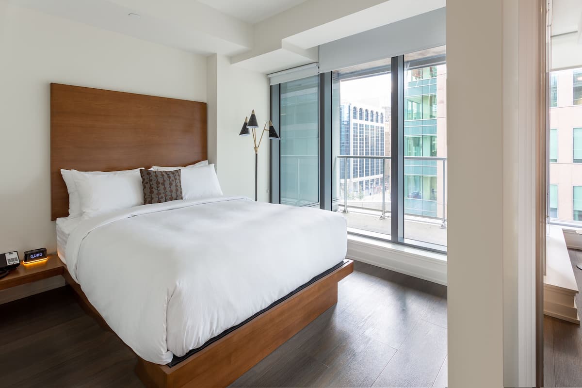 Une chambre d’hôtel moderne dotée d’un grand lit, d’une table de chevet avec une lampe et offrant une vue sur la ville grâce à de grandes fenêtres du sol au plafond.