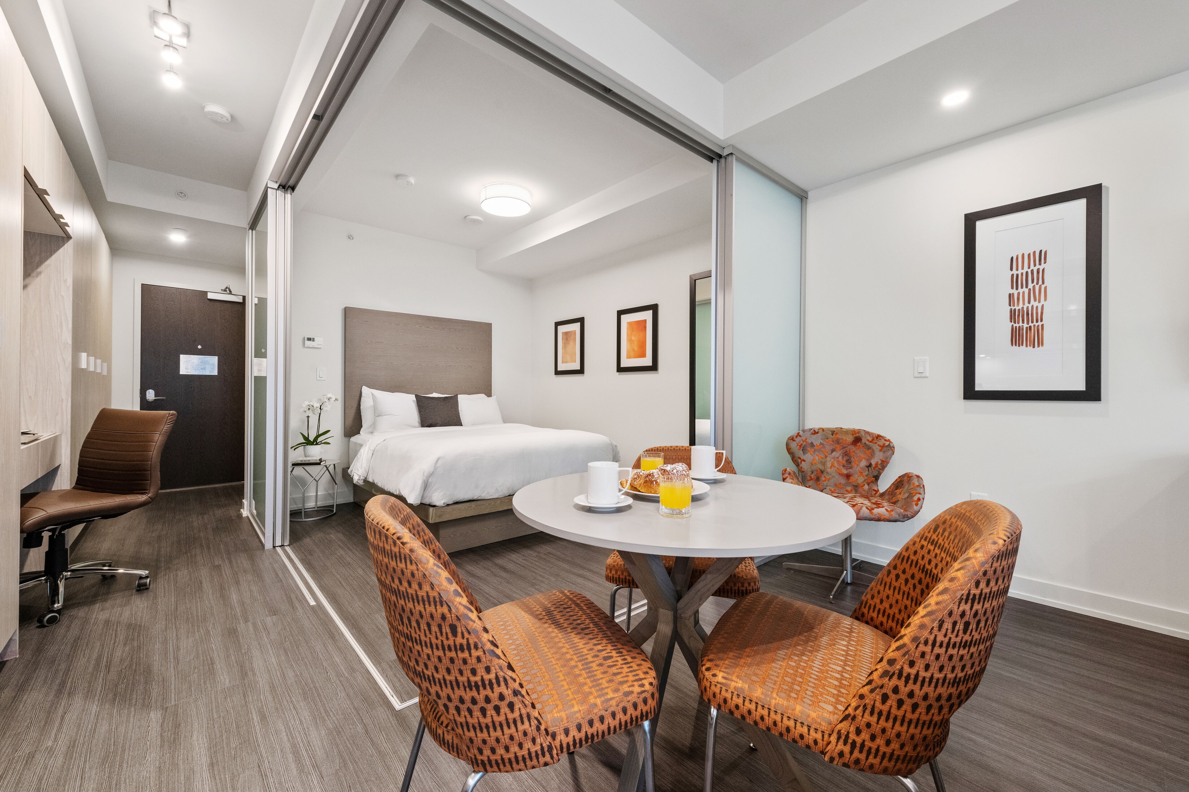 A modern hotel room with a bed, a dining area with chairs, a small table with a fruit bowl, and a wall-mounted artwork.