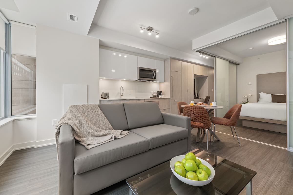 Une chambre d’hôtel moderne avec un canapé gris, un coin repas comprenant une table et des chaises, un coin cuisine équipé d’un four à micro-ondes et un lit avec une tête de lit.