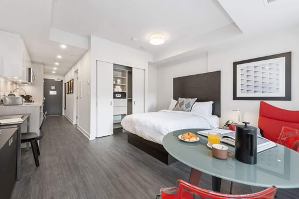 Une chambre d’hôtel moderne avec un lit, une table en verre avec un plateau de petit-déjeuner, une chaise rouge et un coin cuisine.