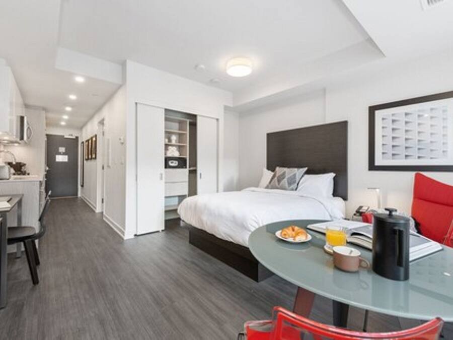 Habitación de hotel moderna con cama, una mesa de vidrio con desayuno, una silla roja y una kitchenette.