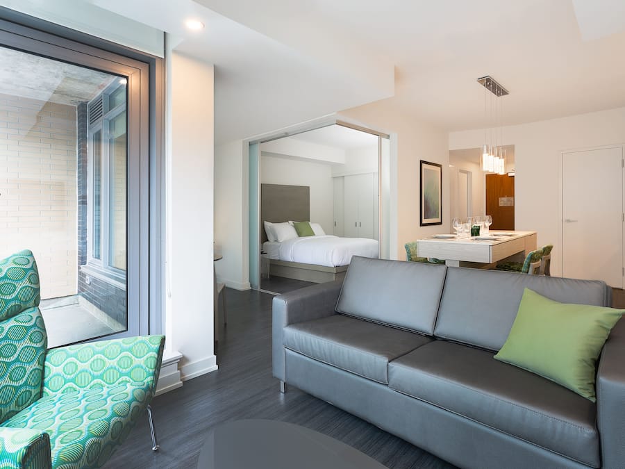 Une chambre d’hôtel moderne avec un canapé gris, un fauteuil à motifs verts, un lit avec une literie blanche, une salle de bain avec un miroir et un coin repas avec une table et des chaises.