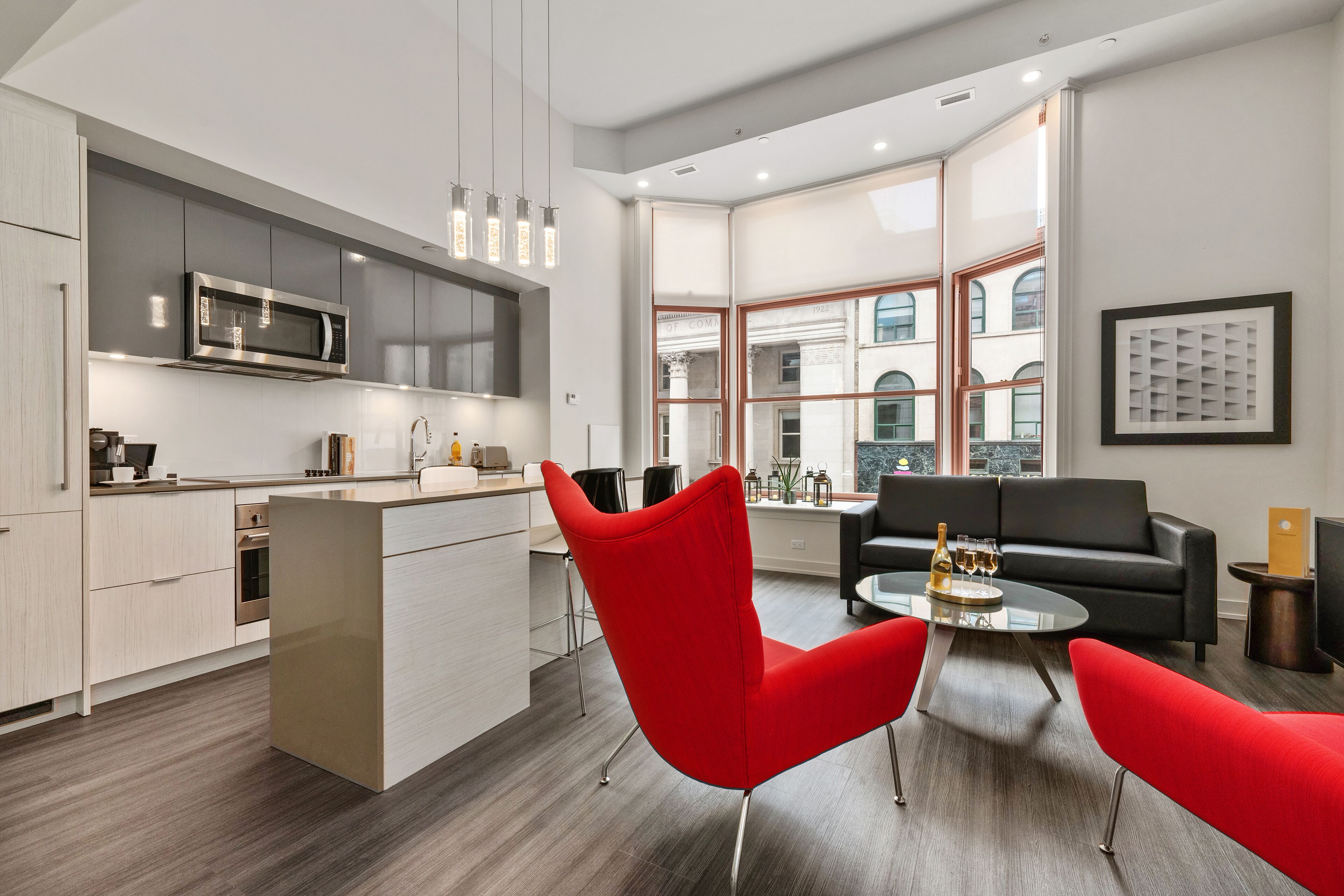 Une cuisine moderne avec un îlot central, un coin repas avec des chaises rouges et une vue sur un immeuble à travers de grandes fenêtres.