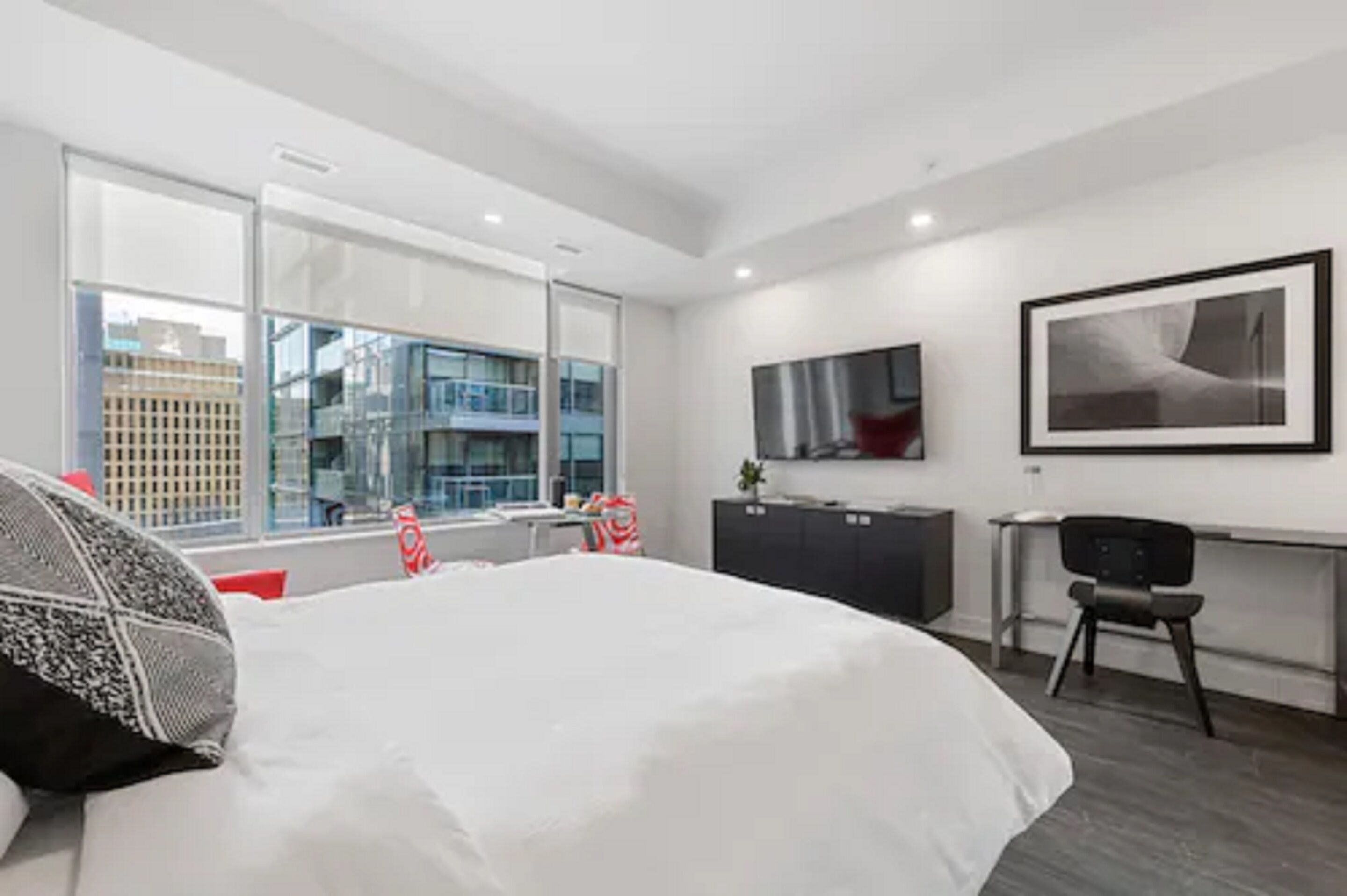 A modern hotel room with a large bed, a desk, a chair, a TV, and a window with a city view.