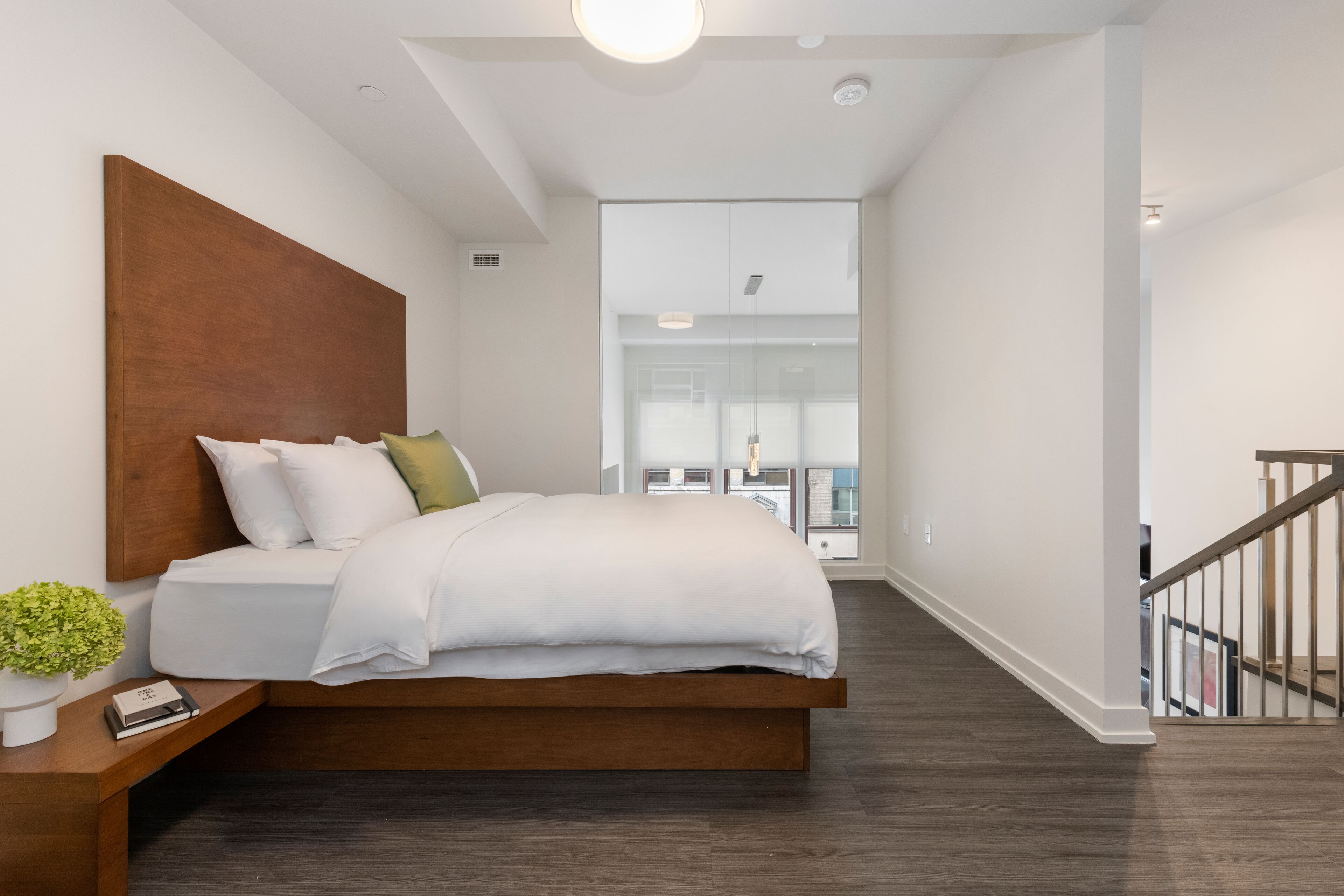 Quarto moderno com cabeceira de madeira, cama grande com roupa de cama branca, mesa de cabeceira com uma planta e vista para uma varanda através de uma porta de vidro.