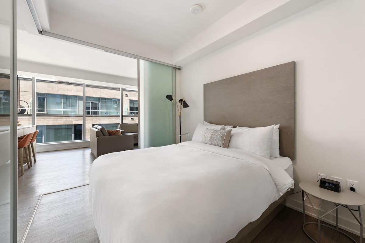 Une chambre d’hôtel moderne dotée d’un grand lit, d’une table de chevet avec une horloge et offrant une vue sur la ville grâce à de grandes fenêtres.