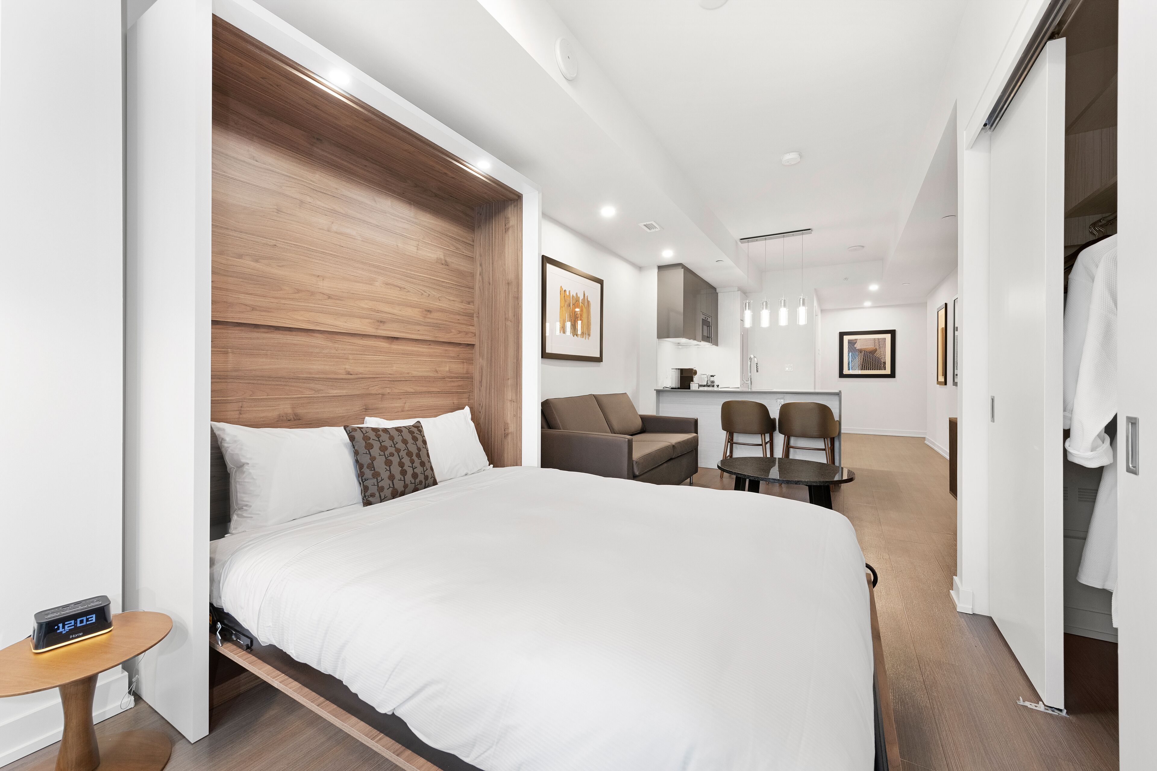 A modern hotel room with a large bed, a wooden headboard, a small table with a clock, a sofa, and a desk with chairs.
