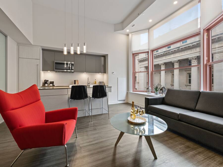 Una sala de estar moderna con una butaca roja, un sofá gris y una mesa de centro de vidrio con una botella y vasos. Se ve una cocina al fondo con un ventanal que da a un edificio.