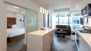A modern hotel room with a bed, a kitchenette, a sofa, and a dining area.