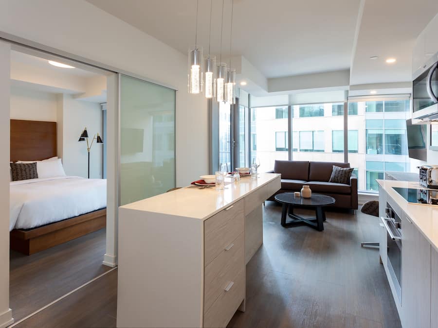 A modern hotel room with a bed, a kitchenette, a sofa, and a dining area.