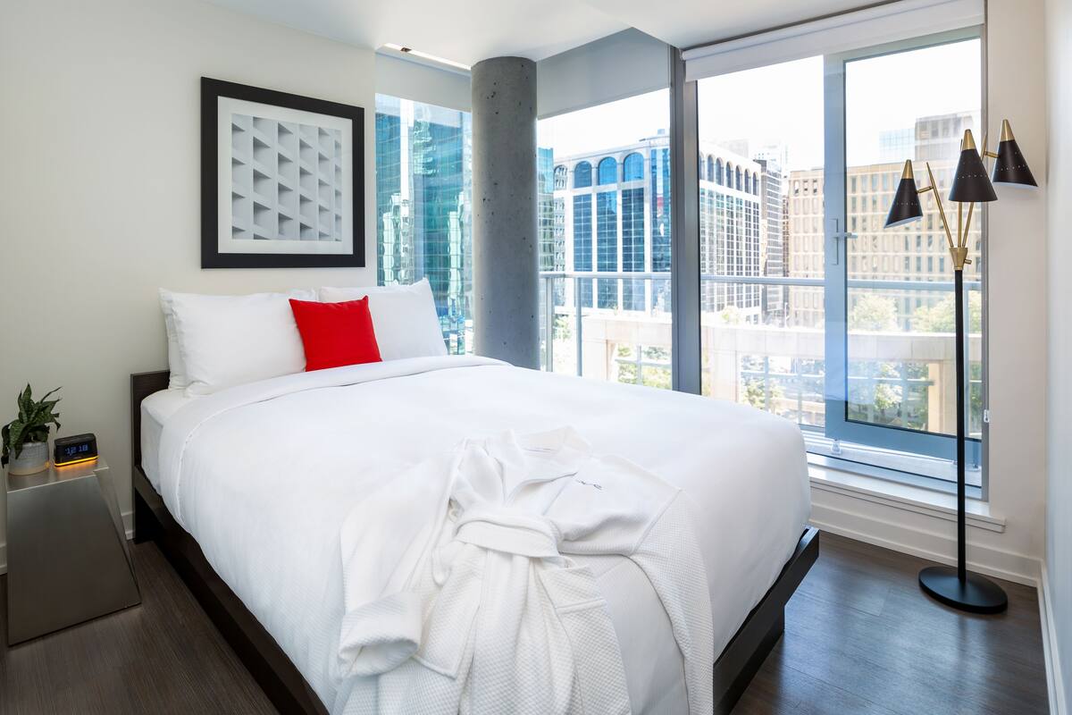 Une chambre d’hôtel moderne avec un grand lit, un oreiller rouge, un peignoir blanc, une horloge numérique et une vue sur la ville à travers de grandes fenêtres.