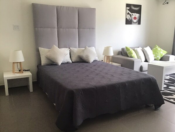 Ein Schlafzimmer mit einem großen Bett, einem grauen, gepolsterten Kopfteil, mehreren Kissen, einem Nachttisch mit Lampe sowie einem Sofa mit Kissen.