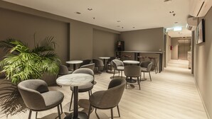 Un área de descanso moderna con mesas y sillas redondas, una planta grande en maceta y una zona de bar al fondo.