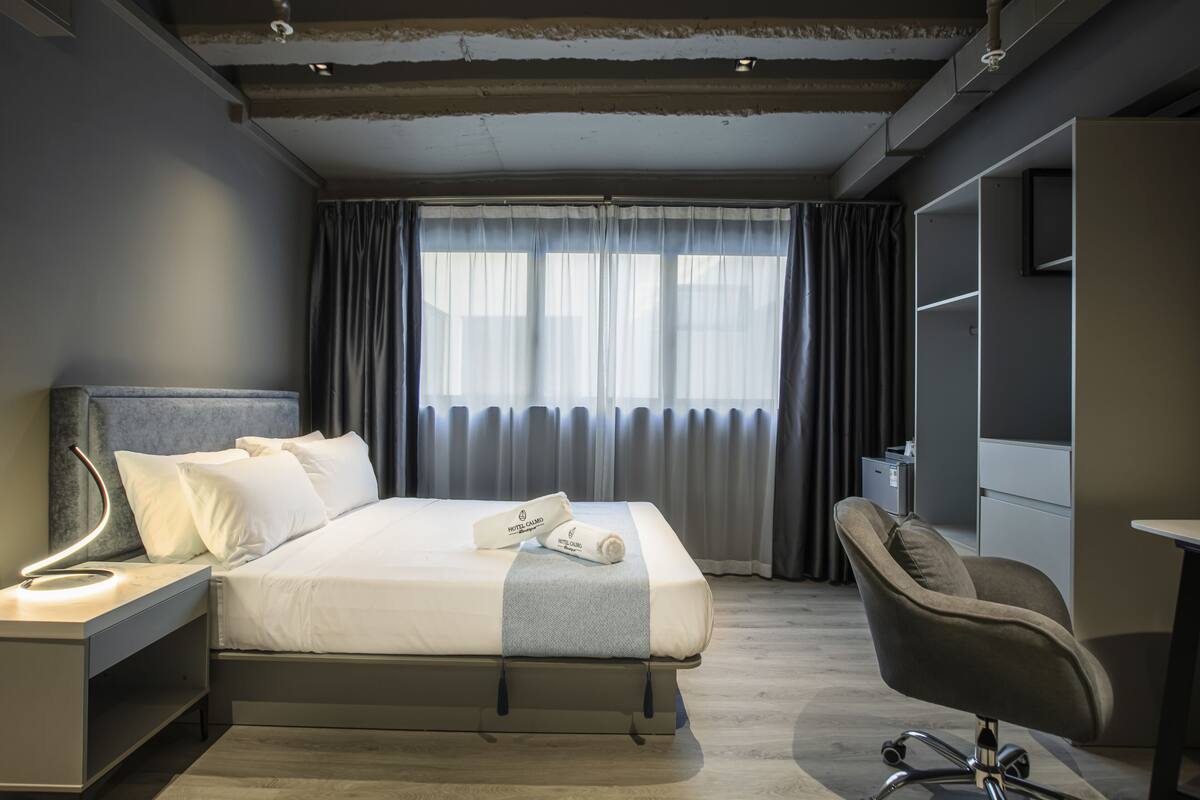 A modern hotel room with a neatly made bed, a desk with a lamp, a chair, and a large window with curtains.