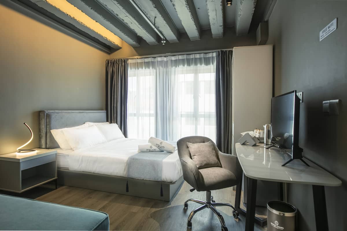 A hotel room with a bed, a desk, a chair, a TV, and a nightstand with a lamp.