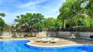A swimming pool with clear blue water, surrounded by a white fence and trees. There are white lounge chairs along the edge of the pool. - Quiet apartment near the sea (Salou)