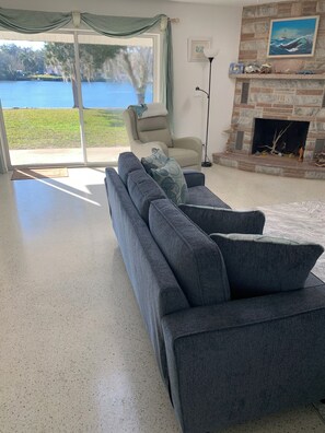 Una sala de estar con un sofá gris, un sillón color beige, una chimenea y vistas a un lago a través de un ventanal corredero.