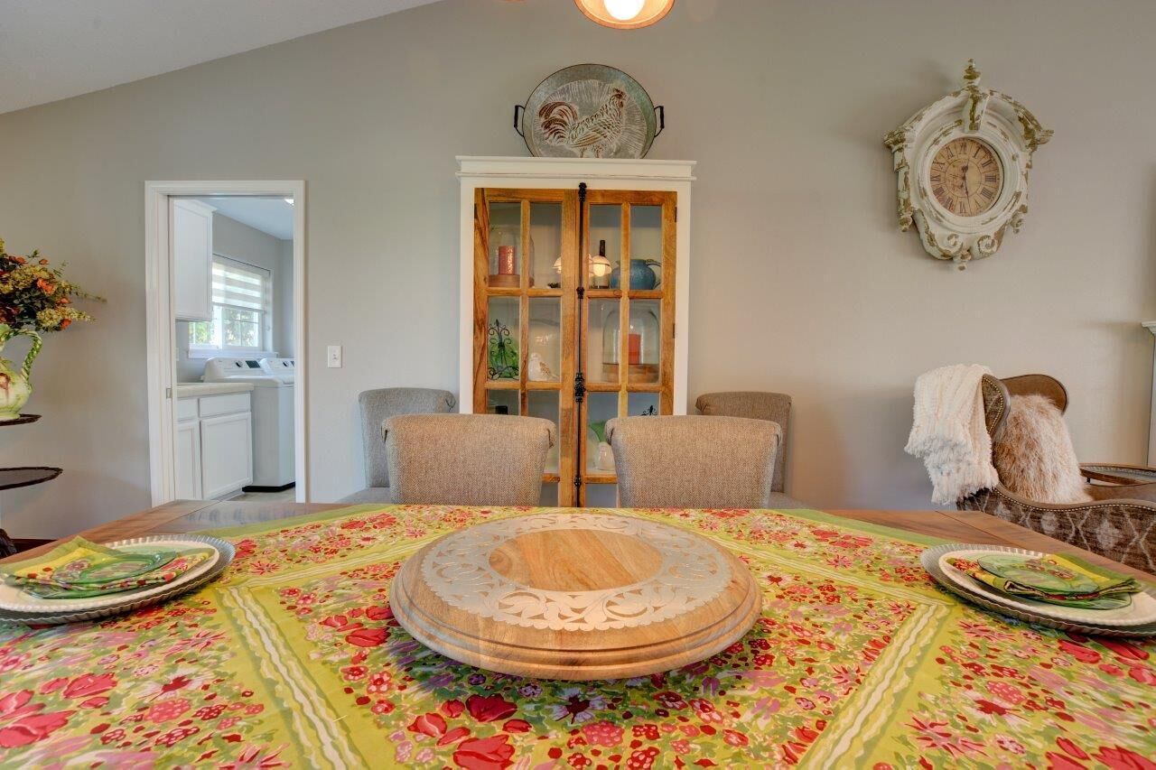 Un comedor con una mesa redonda, sillas y un centro de mesa decorativo. Se observa un aparador con puertas de cristal al fondo, así como un reloj de pared.