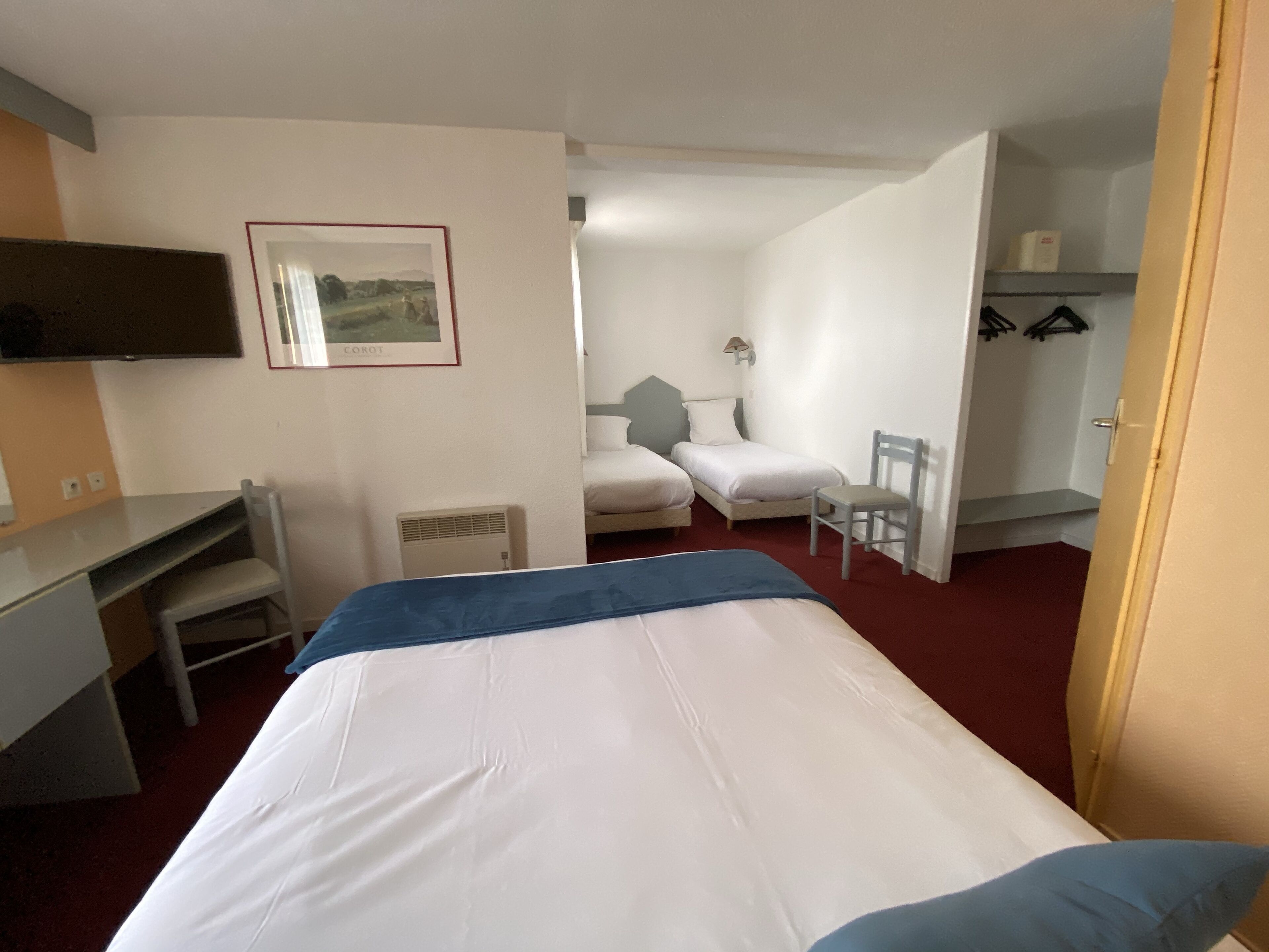 Une chambre d’hôtel avec un lit, un bureau, une chaise et une petite table. Il y a un tableau au mur et une lampe sur le bureau.