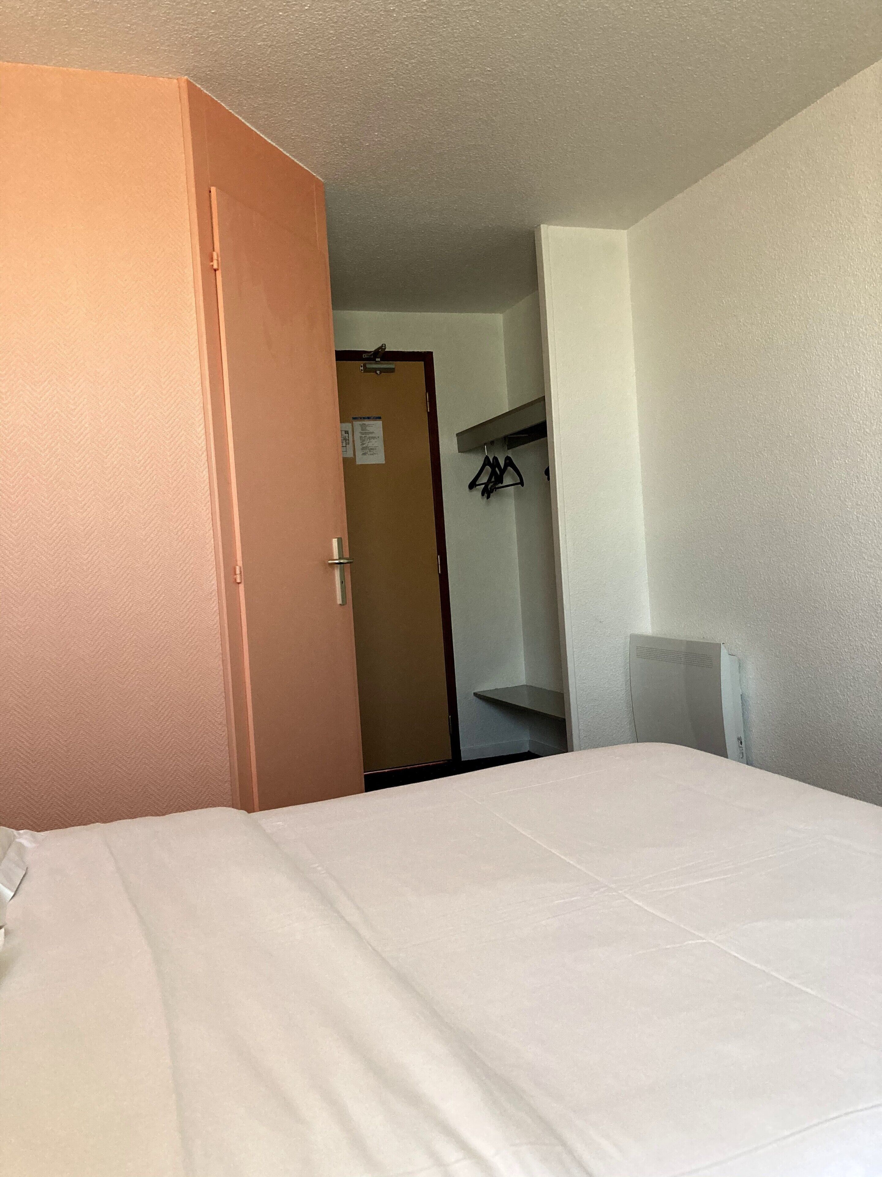 A hotel room with a bed, a wardrobe, and a door leading to a hallway with a coat rack.