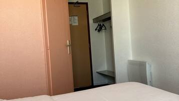 A hotel room with a bed, a wardrobe, and a door leading to a hallway with a coat rack.